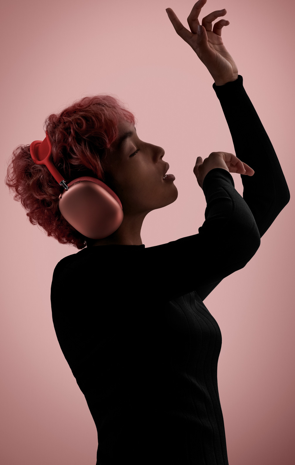 Bočni prikaz osobe u pokretu, zatvorenih očiju i podignutih ruku, koja nosi ružičaste slušalice AirPods Max.
