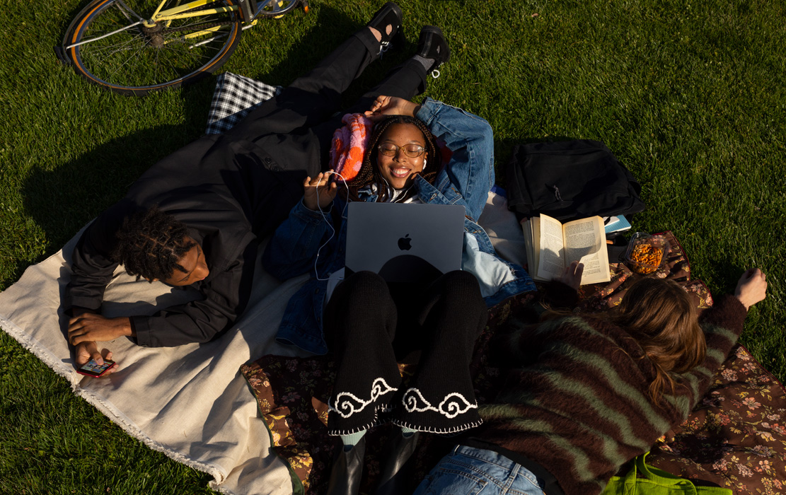 Three university students lie on a blanket in the park. One student has an iPhone. One student has a MacBook Air and Apple headphones. One student has a paperback book.