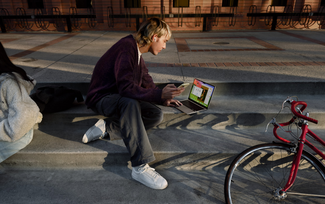 A university student sitting on steps with a bicycle using an iPhone and MacBook